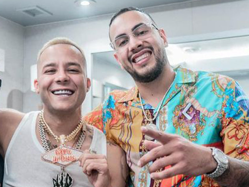 Dos hombres sonrientes luciendo cadenas de oro con colgantes grandes y un reloj plateado con incrustaciones.